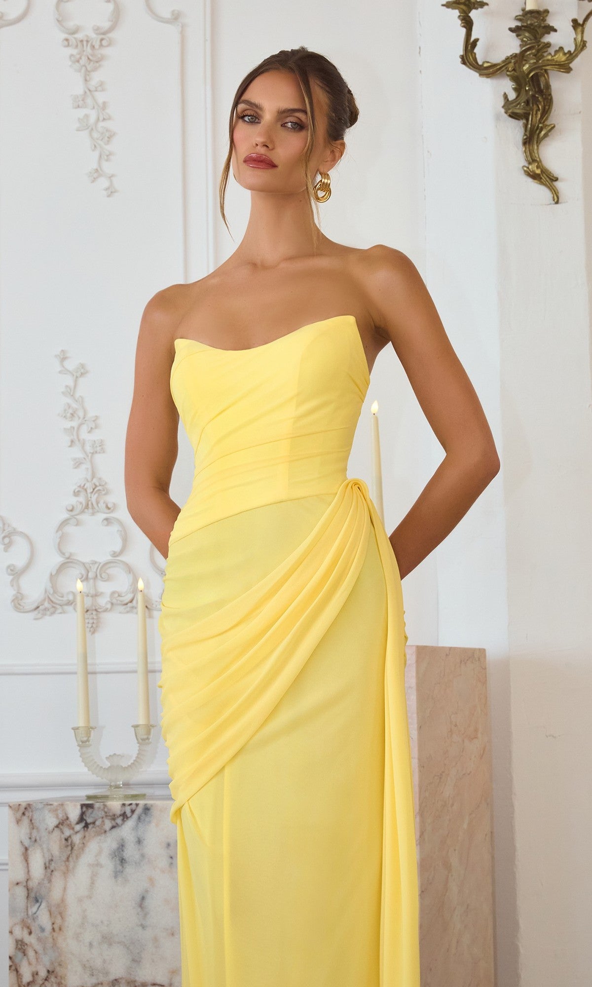 A woman wearing the Amoris T726 Long Prom Dress, featuring a strapless chiffon design with draped details and a lace-up back, stands indoors by a candelabra and marble surface.